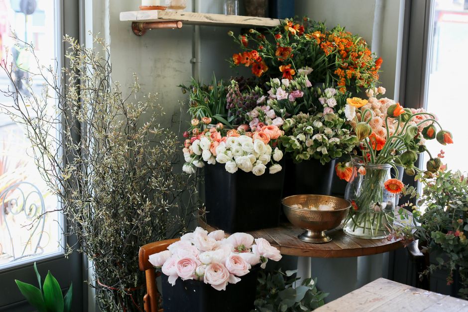 An indoor display of freshly cut flowers arranged in black, clear, and metallic vessels on a round wooden table near a window, with natural light illuminating the vibrant bouquet of pink and white ros