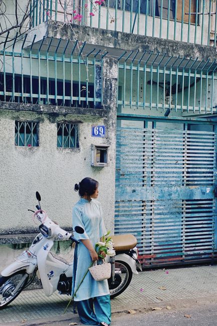 A woman dressed in traditional light blue and darker blue clothing stands on a sidewalk near a white scooter, holding a bouquet of fresh flowers wrapped in light paper. She faces slightly to the left,